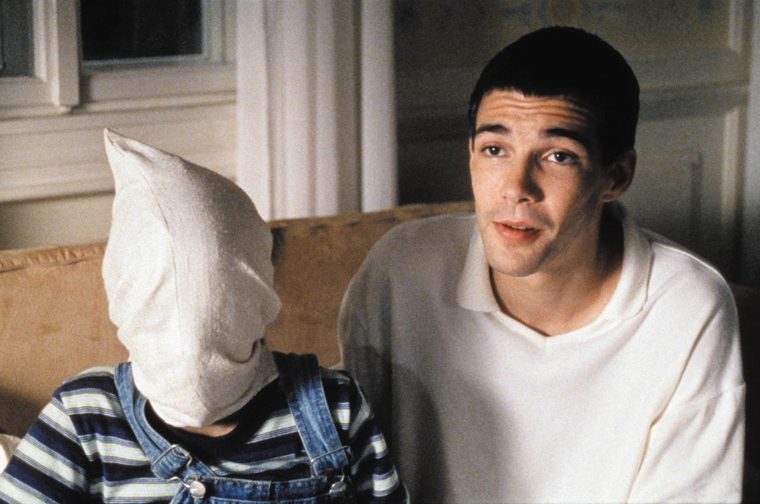 A young man in a white polo shirt sits on a sofa. Next to him is a young boy wearing a stripy shirt and blue dungarees.. He has a white bag over his head.