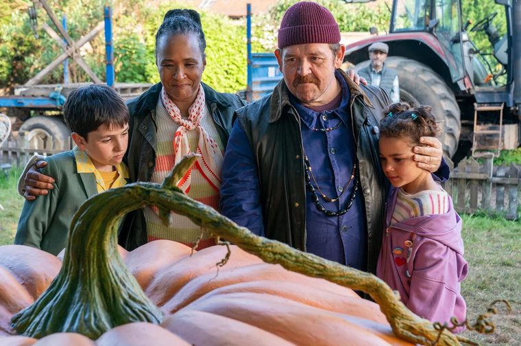A heartwarming scene from a community event centered around an impressively large pumpkin. The image shows a family of four - two adults and two children - gathered around what looks like a giant competition-sized pumpkin, the kind you might see at a harvest festival or pumpkin-growing competition. The setting has a lovely rural or farm atmosphere, with a tractor visible in the background, wooden structures, and autumn foliage. Everyone seems engaged and interested in examining this enormous gourd, which has that characteristic pale orange color and thick, twisted stem that giant pumpkins develop.