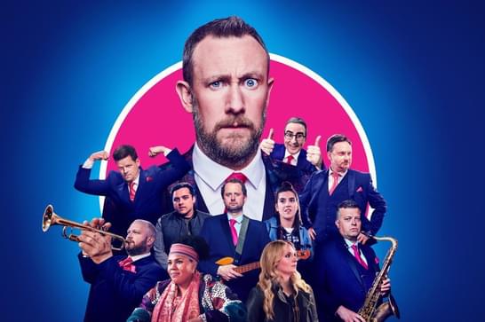A collection of men in dark blue suits can be senn crowded around the giant head of Alex Horne, who has a beard and a red tie on. Some of the men have musical instruments, including a saxophone, and there are three women at the front.