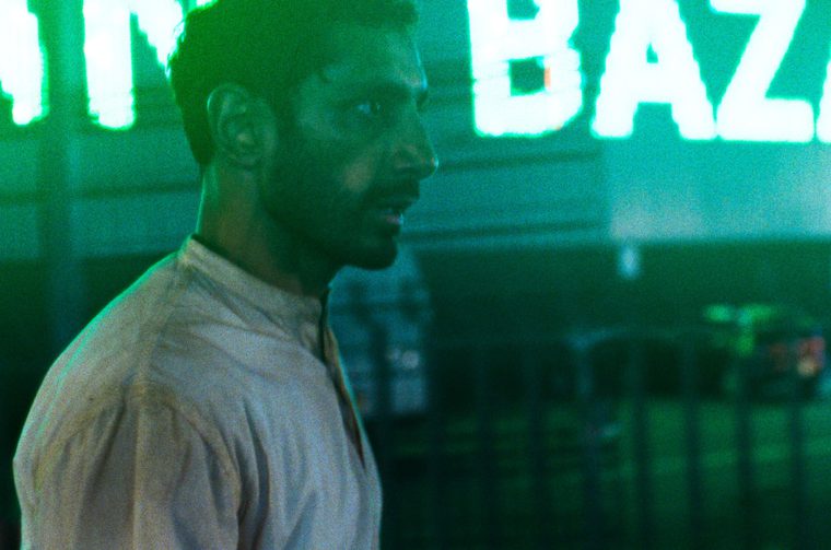 A fuzzy image of a South Asian man (Riz Ahmed) in his forties as he walks along a street in London, giant neon lettering behind him that start to spell out the word 'Bazaar.'