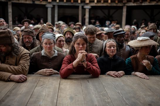 The image shows a young woman in the foreground, dressed in a burgundy or rust-colored garment, with her hands clasped together in what appears to be prayer or anguish. She's positioned at what looks like a wooden barrier or railing, with a large crowd of period-dressed townspeople gathered behind her, all wearing earth-toned clothing and various head coverings typical of peasants and commoners from that era.