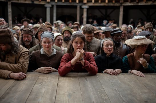 The image shows a young woman in the foreground, dressed in a burgundy or rust-colored garment, with her hands clasped together in what appears to be prayer or anguish. She's positioned at what looks like a wooden barrier or railing, with a large crowd of period-dressed townspeople gathered behind her, all wearing earth-toned clothing and various head coverings typical of peasants and commoners from that era.