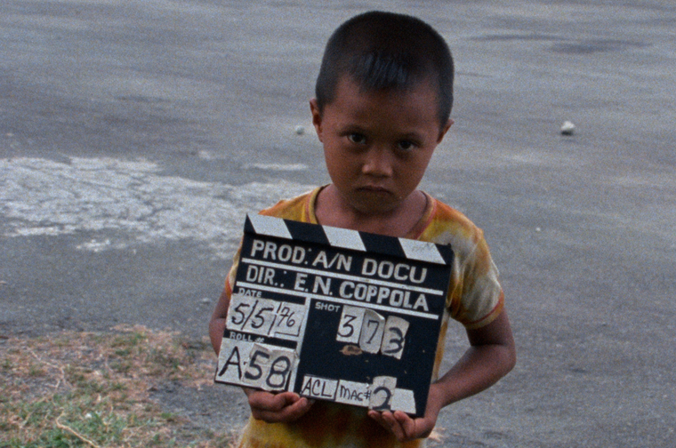 Small child holding a clapperboard that reads 'Prod: A/N Docu, DIR.: E.N. Coppola, Date: 5/5/76' The image was taken on the set of Francis Ford Coppola's Apocalypse Now.