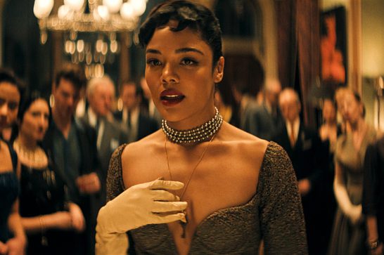 We see an elegantly dressed woman in a formal setting with ornate chandeliers and a crowd in the background. The scene has a rich, cinematic quality with warm golden tones.