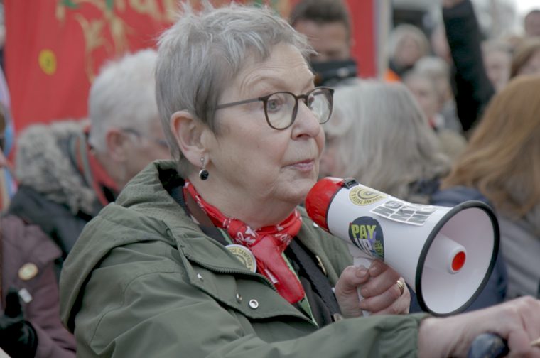 We see an older woman with short gray hair and glasses at what appears to be a protest or demonstration. She's wearing an olive green jacket with a red bandana around her neck and is holding a megaphone decorated with stickers including one that says "PAY" and another that appears to say "FIGHT FOR 15" (a campaign related to minimum wage advocacy). There are other people visible in the background, and red banners or flags can be seen.
