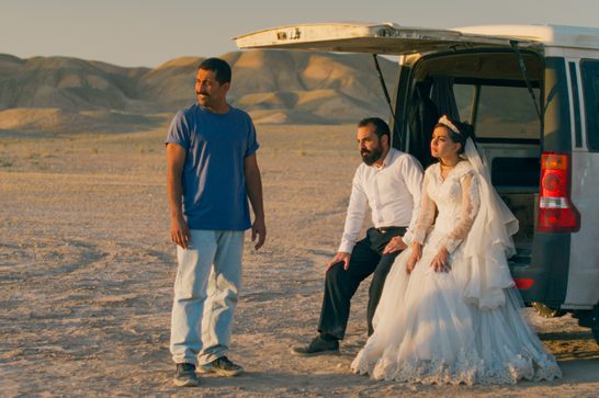 The image shows three people near a vehicle with its back door open: a man in casual attire (blue t-shirt and light pants) standing to the left, and what appears to be a couple in more formal dress sitting in the vehicle's cargo area. The person on the right is wearing an elaborate white dress that could be a wedding gown or formal costume piece. The setting sun creates warm, golden lighting across the arid terrain, with dramatic hills or badlands visible in the background.