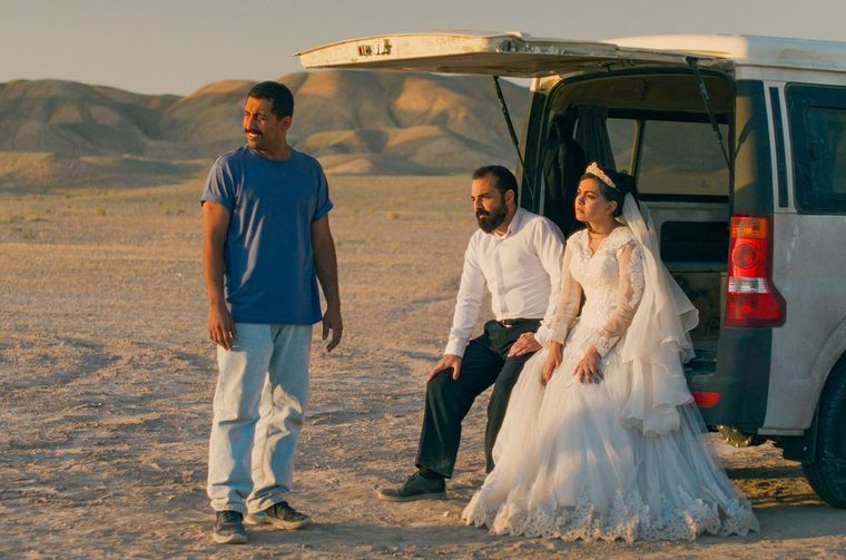 Three people in a desert landscape at golden hour - one man in a blue shirt standing, and a couple in wedding attire sitting at the back of an open SUV with mountains in the background.