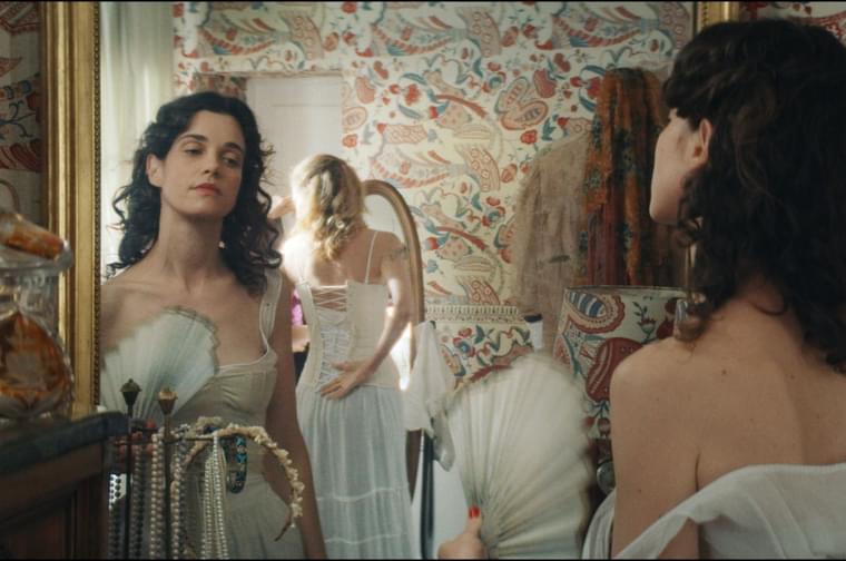 A woman in her thirties stands in front of a mirror, looking at her own reflection. She is wearing a white dress and holding a white fan. Behind her is another woman whose face is turned away from the camera, also looking into a mirror and doing up the back of her own white dress.