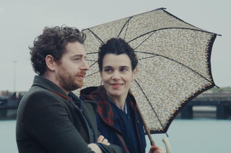 A man and a woman in their thirties walk alongside an embankment. The man on the left has a beard and has his arms crossed, whilst the woman on the right looks adoringly at him, holding an open umbrella that rests on herleft shoulder.