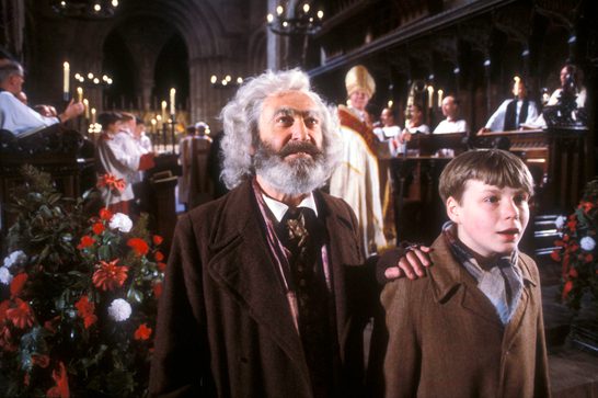 A screen shot of the TV series of Box Delights showing an elderly man with a bushy grey beard standing behind a young boy in a brown jumper, his left hand on the boy's right shoulder. They are in a church and behind them are a choir, a bishop and numerous lit candles.
