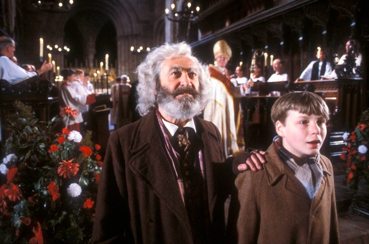 A screen shot of the TV series of Box Delights showing an elderly man with a bushy grey beard standing behind a young boy in a brown jumper, his left hand on the boy's right shoulder. They are in a church and behind them are a choir, a bishop and numerous lit candles.