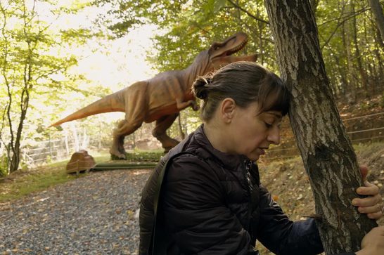 The image shows a woman hiding behind a tree while a large animatronic theropod dinosaur (possibly a T-Rex or similar species) looms behind them in the background. The women has her forehead resting on the tree trunk and her eyes closed.
