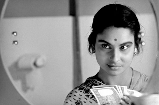 A black and white photo of a young woman with traditional South Asian styling - her hair is pulled back, she's wearing a bindi, and has decorative earrings. She's captured both directly in the frame and reflected in what appears to be a round mirror on the left side, creating an artistic dual perspective. She's holding what looks like a stack of cards or photographs in her hands, and she has a gentle, contemplative expression.