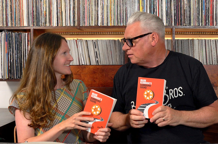 Photography of a man and woman both holding the same book and looking at each other. The book has an orange cover and piano keys leading to a film reel. Behind them is a wall of vinyl record.
