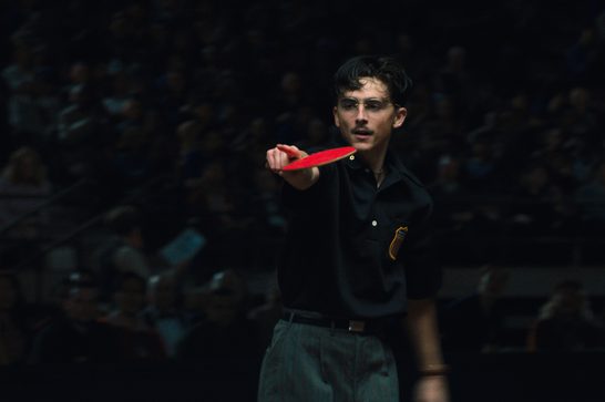 A young male table tennis player with a moustache and glasses is captured mid-action during what appears to be a competitive match. The dramatic lighting creates a cinematic quality, with the player emerging from a dark background while intensely focused on the game. He's wearing a black athletic shirt and is shown in a ready position, holding a red paddle extended forward.