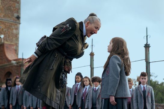 We see a stern headmistress or authority figure in an olive-green military-style coat adorned with various tools and implements, looming intimidatingly over a young schoolgirl in a grey uniform. The setting appears to be a schoolyard with other uniformed students in the background.