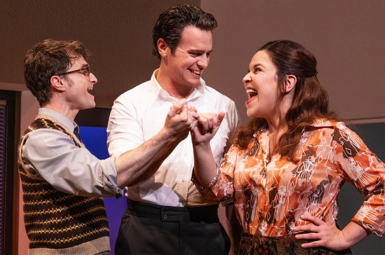 Two men and one woman are laughing as they all link their little fingers in a show of friendship. The man on the left wears glasses and a stripey woollen vest. The man in the middle wears a white shirt and black trousers. The woman on the right is wearing an orange and brown blouse.