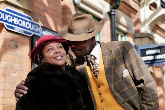 An older Black man wearing a trilby, brown suit and yellow waistcoat hugs a smiling Black woman with his right hand. She is wearing a black coat and a pink hat. They are standing in a train station, the word Loughborough Central visible on a blue and white sign behind them.