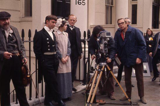 A behind-the-scenes photograph from a TV production, likely from the 1970s or 1980s based on the film equipment and overall aesthetic. The scene shows actors in period costume - what appears to be Victorian or Edwardian era clothing - standing outside a Georgian-style building with the number "165" visible above the door. The costumes suggest this might be from a period drama or historical film. There's a woman in a maid's uniform with a white cap, men in formal Victorian attire including one in what looks like a naval uniform, and others in period-appropriate clothing.
