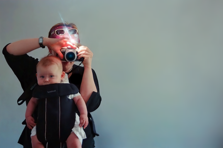 A photograph of a woman taking a picture of her reflection with a film camera. The camera is covering her face. She has a baby in a baby carrier strapped to her chest.