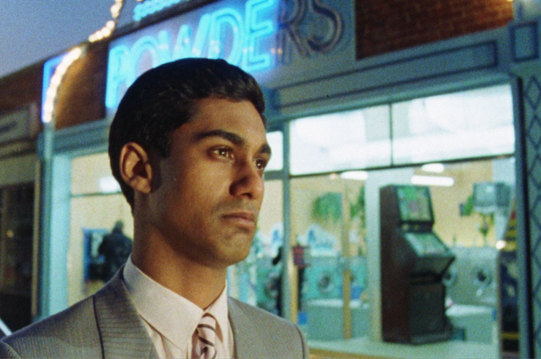 Close up of a young Indian man stood outside of the laundrette he owns. He is wearing a grey suit with a black and white tie.