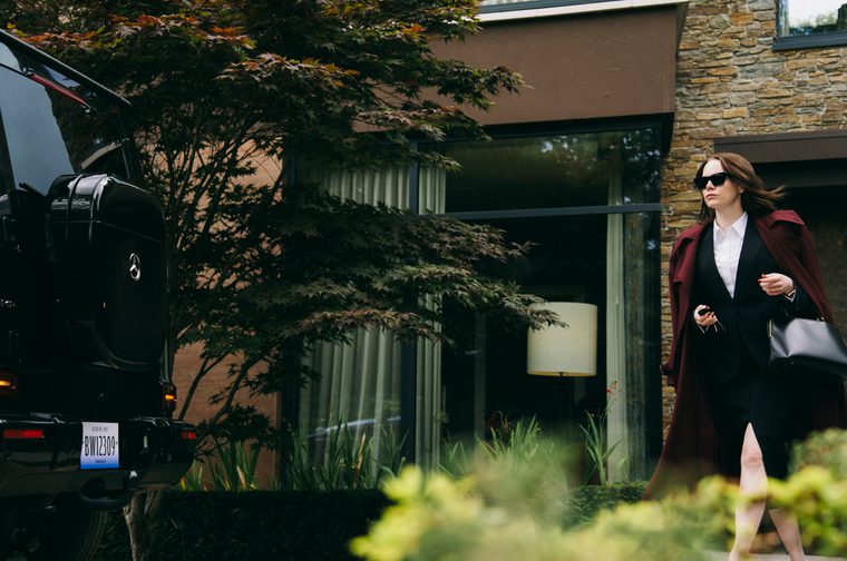 Woman in sunglasses, black blazer, and burgundy coat walking past a modern stone and glass house with a black SUV parked outside.