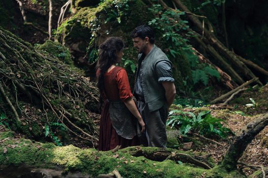 A woman in a red dress and apron stands facing a man in a gray shirt in a lush, moss-covered forest setting. They appear to be having an intimate conversation among fallen logs, ferns, and dense green vegetation. The natural lighting filters through the trees, creating a romantic and atmospheric woodland scene.