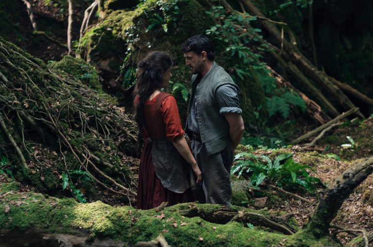 A woman in a red dress and apron stands facing a man in a gray shirt in a lush, moss-covered forest setting. They appear to be having an intimate conversation among fallen logs, ferns, and dense green vegetation. The natural lighting filters through the trees, creating a romantic and atmospheric woodland scene.