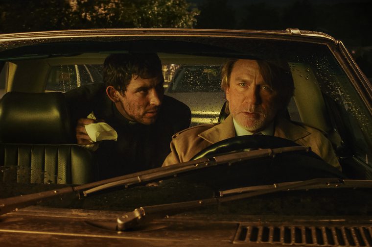 Two men sitting in the front seats of a vintage car at night. The driver (Daniel Craig), an man with graying hair and beard wearing a tan coat, grips the steering wheel while leooking ahead with a serious expression. In the passenger seat, a younger man (Josh O’Connor) with dark hair wearing a dark jacket holds a white paper bag. The scene is dimly lit with warm, golden lighting filtering through the car windows, creating a moody, film noir atmosphere. Rain droplets are visible on the windshield.