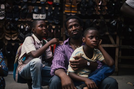 Three young men from Lagos sit closely together on what appears to be a step or ledge in front of a bustling shoe store. The father in the center wears a purple striped shirt and has his arms around his two sons - one wearing a light-colored collared shirt on the left, and another in a yellow and blue striped shirt on the right. Behind them is shelves displaying shoes.