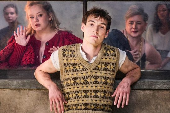 The image shows a young man in the foreground wearing a distinctive tan/beige patterned sweater vest over a white shirt, sitting in what looks like a contemplative or somewhat uncertain pose. Behind him, visible through what appears to be a window or glass partition, are several other people including a woman in a striking red/burgundy textured jacket on the left.