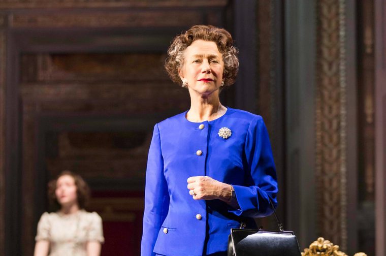 Queen Elizabeth II (played by Helen Mirren) stares out into the distance, wearing a bright blue dress with a pearl brooch pinned on it just underneath her left shoulder. She holds a handbag on her left arm. We can see a young girl in the background, blurry but noticeably wearing a frilly white dress.