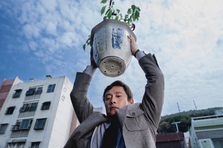 An Asian man in his forties holds up a large ceramic or porcelain pot that's being creatively repurposed as a planter, with a small tree growing from it. The pot appears to be a traditional Asian-style container, possibly a vintage piece, with blue decorative elements visible on its white surface. The drainage holes at the bottom are clearly visible. The photograph is taken from a low angle, making the person and pot appear dramatic against the cloudy sky. In the background, you can see a multi-story residential building and some hillside greenery, suggesting an urban Asian setting.