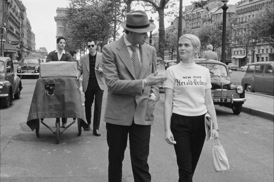The black and white image shows a film production on location in Paris, with the Arc de Triomphe visible in the background. The scene depicts a director (in the suit and fedora) working with an actress wearing a "New York Herald Tribune" sweater - a distinctive costume choice that became iconic in French cinema of this era. Behind them, crew members are setting up equipment, including what looks like a camera on a tripod under a cloth covering.