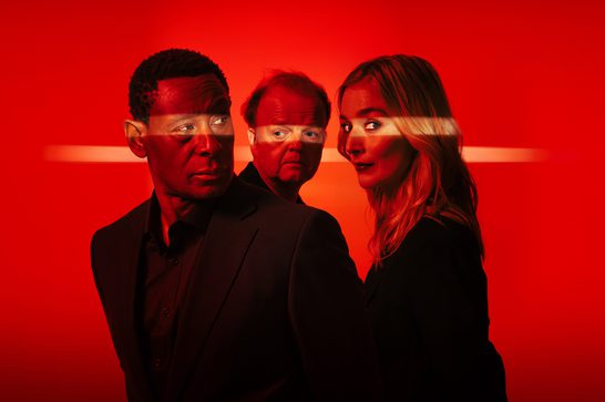 We see a heavily stylized image of an older Black man (David Harewood), a younger white woman with blonde hair (Caitlin Fitzgerald) and a balding older white man (Toby Jones) standing next to each other, all wearing black and all looking in different directions. The rest of the image is almost enturely red, with a yellow line of light shining horizontally across the eyes of each of the peopl.