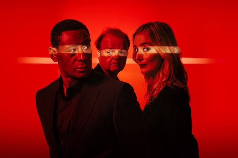 We see a heavily stylized image of an older Black man (David Harewood), a younger white woman with blonde hair (Caitlin Fitzgerald) and a balding older white man (Toby Jones) standing next to each other, all wearing black and all looking in different directions. The rest of the image is almost enturely red, with a yellow line of light shining horizontally across the eyes of each of the peopl.