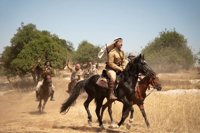 A photo depicting armed horsemen in Middle Eastern military attire from the early 20th century. The scene shows several riders on horseback moving through an arid landscape with olive trees in the background. The lead figure on the black horse wears a tan/khaki military uniform with a traditional keffiyeh (head covering), and carries what appears to be a rifle. The group behind him also sports similar period military dress and headwear.