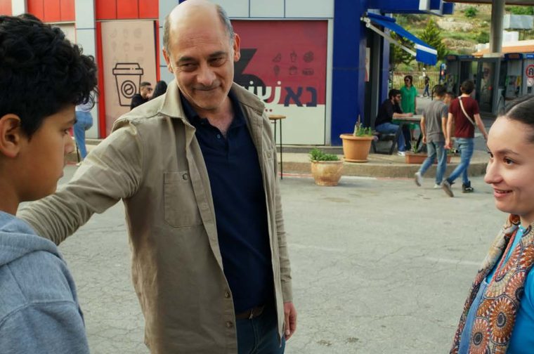 three people having a conversation at what looks like a public transit area or bus station. There's a boy on the left, a middle-aged man in the center wearing a tan jacket, and a woman on the right wearing a blue shirt and patterned scarf. The setting appears to be on a Middle Eastern street, with other people passing by.