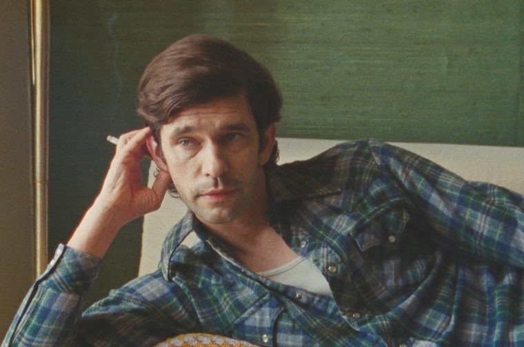 A young man (Ben Whishaw) in a blue and green plaid flannel shirt, positioned in what looks like a thoughtfully composed shot. The lighting is soft and natural, coming from what seems to be a window, creating a gentle illumination on one side of his face while leaving subtle shadows on the other.