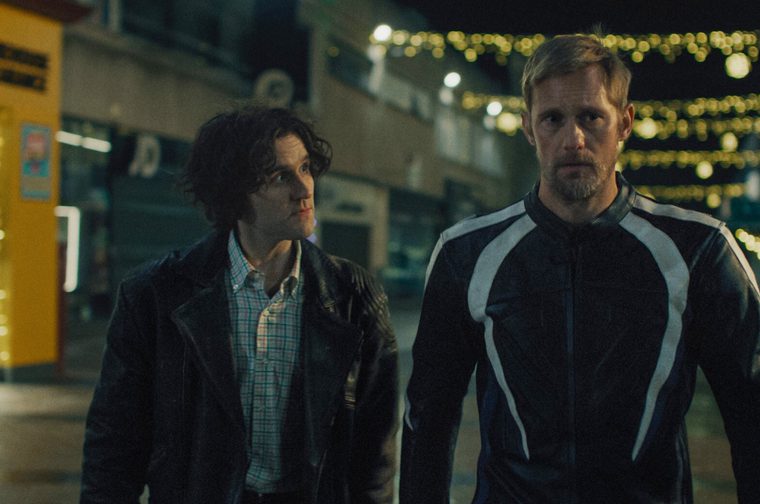 Two men stand together on a nighttime street illuminated by string lights overhead. The younger man on the left wears a dark leather jacket over a checked shirt, while the older man on the right is dressed in a black and grey biker jacket.