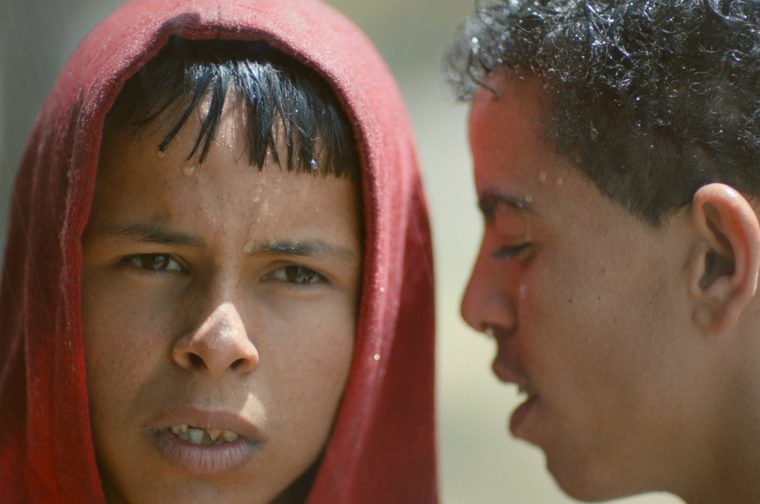 A young Tunisian boy stares just off camera, a red hood over his head. Another Tunisian boy stands next to him, talking into his ear. Both are wet from the pouring rain.