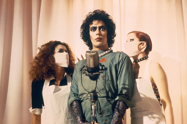 A promotional photo from "The Rocky Horror Picture Show," the iconic 1975 cult musical film. The image shows three people in costume - the central figure is wearing the distinctive makeup and costume associated with Dr. Frank-N-Furter, complete with dramatic eye makeup, curly dark hair, and what appears to be a corset or fitted top. They're positioned at a microphone