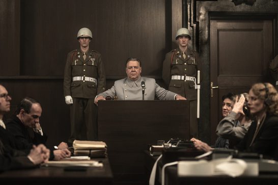 A Nazi commander (Russell Crowe) sits behind a desk with a microphone, staring into the camera. On either side in front of him are various men and women in suits, most wearing headphones. Behind the Nazi commander stand two soldiers wearing grey helmets, standing to attention.