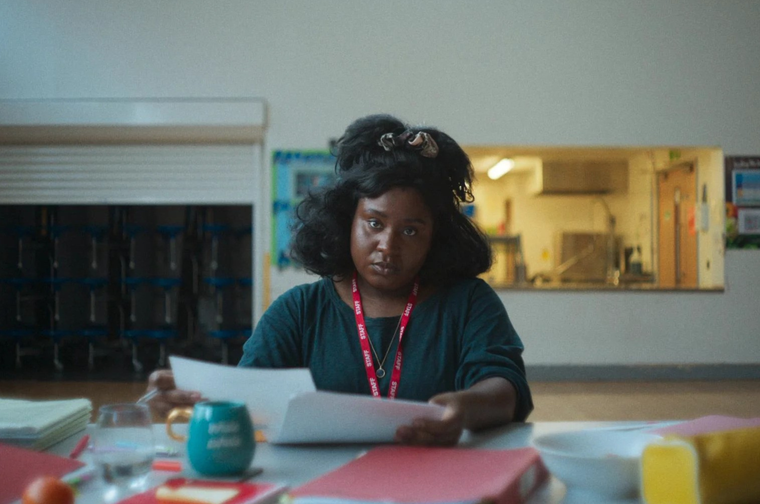 A woman with black hair sits at a table holding papers, wearing a red lanyard and looking directly at the camera.