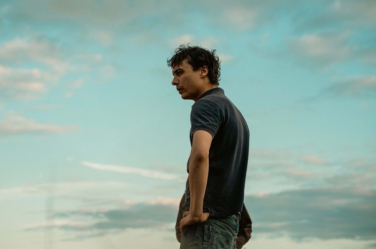 A contemplative portrait of a young man with curly hair wearing a dark t-shirt and jeans, photographed against a beautiful sky with soft, wispy clouds. The image has a cinematic quality with warm, golden lighting that creates a peaceful, reflective mood. The person appears to be looking off into the distance, captured in profile against the expansive sky backdrop.