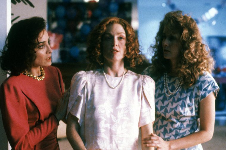 A thirtysomething woman with ginger hair stares off camera, whilst two other women stand either side of her, resting their hands on her arms. They all wear pearl necklaces and are wearing 90s clkothing, with the black haired woman on the left wearing a red dress and the curl blonde haired woman on the right wearing a white dress with blue butterfly patterns..