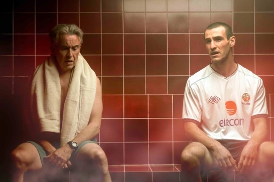 We see a scene from what looks like a sauna or steam room, with two men sitting against a distinctive terracotta-tiled wall. The younger man on the right is wearing an Ireland national team football jersey with "eircom" sponsorship visible on it. The older man on the left has a towel draped around his shoulders.