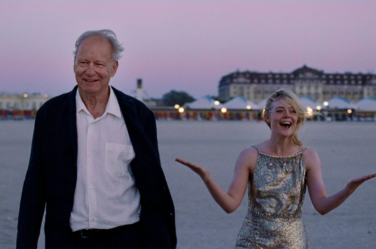 The image captures two characters in an outdoor setting - an older gentleman in casual formal attire (black jacket and white shirt) and a younger woman in a glamorous, sequined sleeveless dress who appears to be expressing joy or excitement with her arms outstretched.
