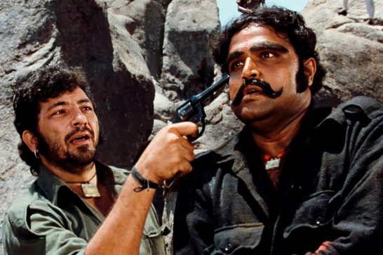 The image shows two of the main characters, Veeru (played by Dharmendra, on the left) and Gabbar Singh (played by Amjad Khan, on the right), in what looks like a tense confrontation in a rocky, mountainous setting.