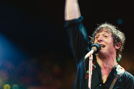 A dynamic concert photograph capturing a musician performing on stage. The image shows a singer with curly hair at a microphone, appearing to be mid-performance with an expressive, energetic delivery. The lighting creates a dramatic atmosphere with effects from stage lights visible in the background - blues, greens, and warm tones creating that classic concert ambiance. There's another figure partially visible in the background, likely another band member. The dark background with colored light spots and the performer's passionate expression really convey the energy and atmosphere of a live music performance.